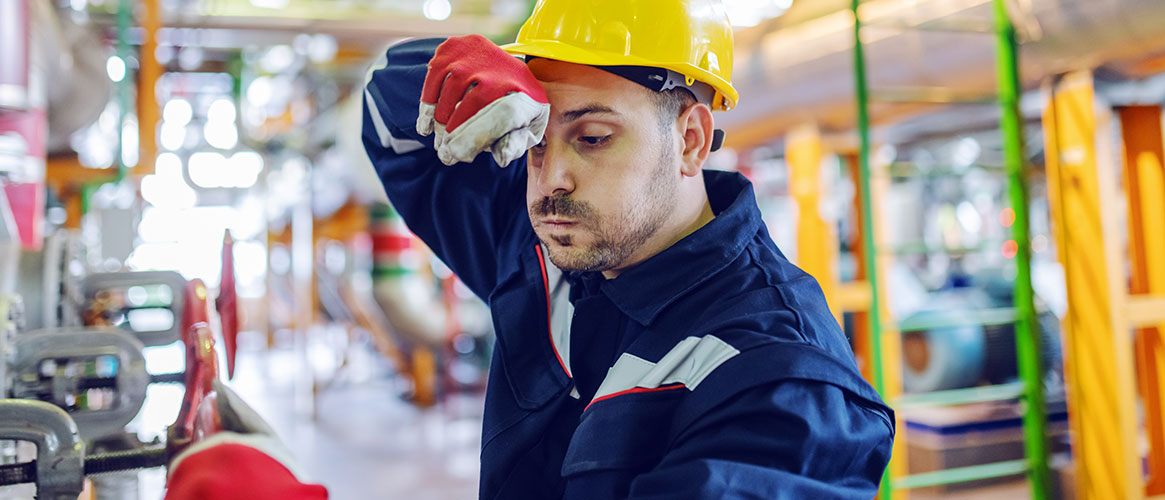 Plant worker in protective suit and helmet trying to screw valve while wiping sweat